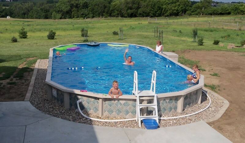 A Doughboy above ground swimming pool in a backyard. There is a family playing in the water with some pool toys scattered throughout.